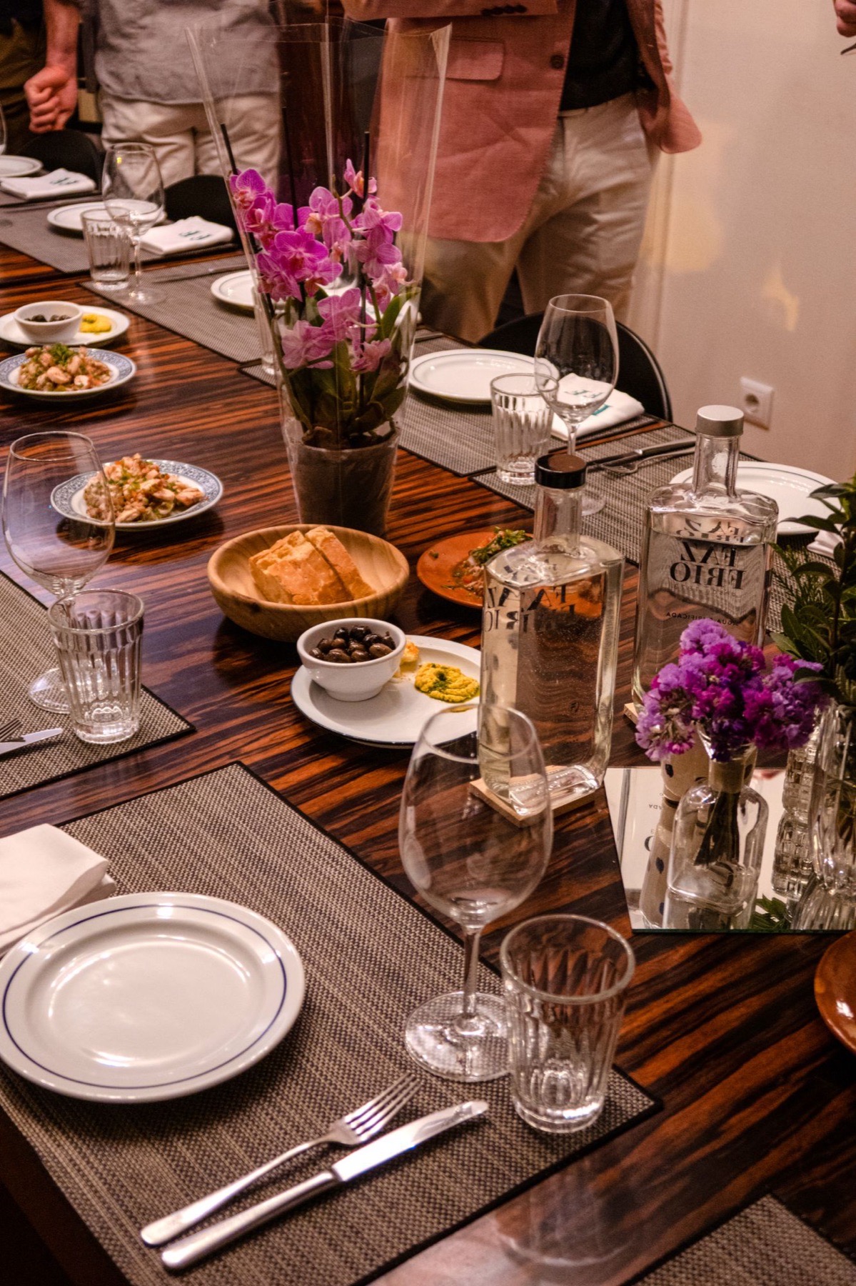 Uma mesa de jantar comprida posta, com pratos, copos e flores, enquanto os convidados se reúnem